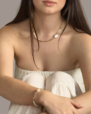 A woman wearing a delicate gold chain necklace with white pearl-like beads and a matching gold bracelet with a pearl accent, styled with a white strapless top against a neutral gray background.