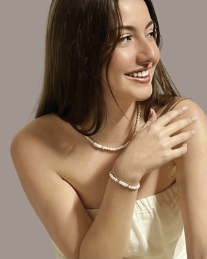 A smiling woman with long brown hair models wearing the Eva Freshwater Pearl Bracelet by CVLCHA, a delicate white pearl necklace styled with modern neutral fashion for effortless everyday elegance.