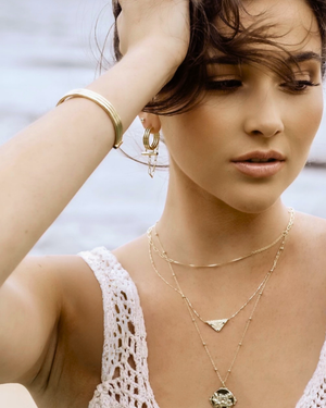 Model Woman looking away while wearing CVLCHA Adira Rod Hoop Earrings with CVLCHA jewellery including a necklace, earrings, and bracelet against blurred background.